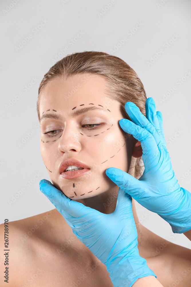 Plastic surgeon touching face of young woman with applied marks on light background