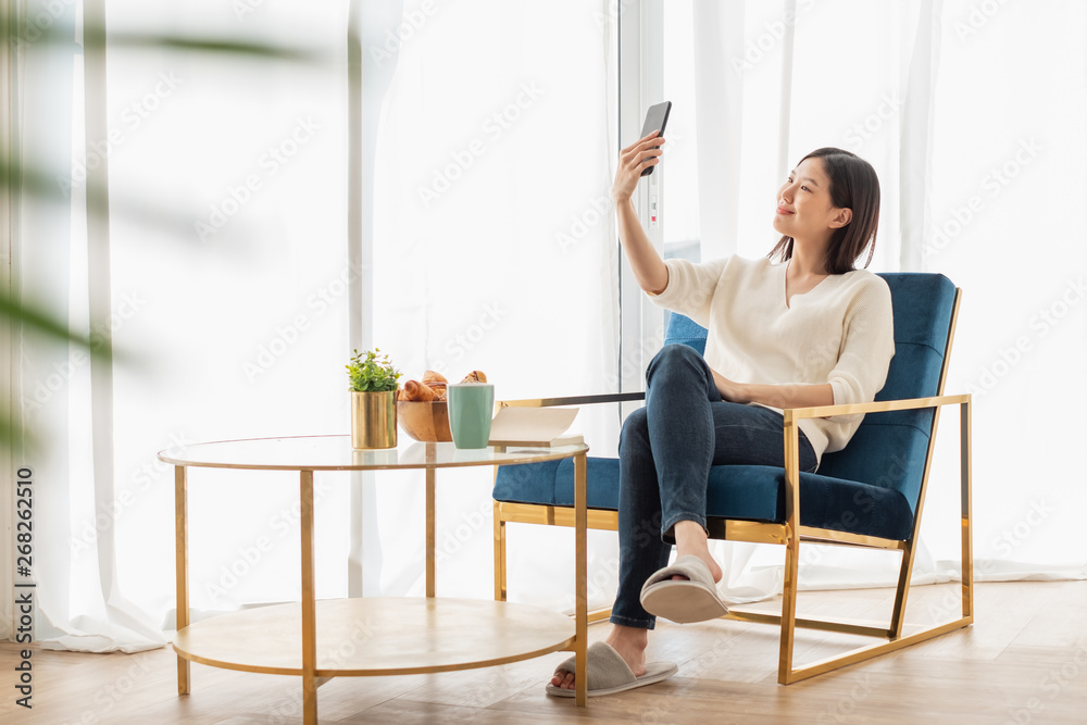 Young beautiful Asian woman relaxing in living room at home, reading books, drinking coffee and usin