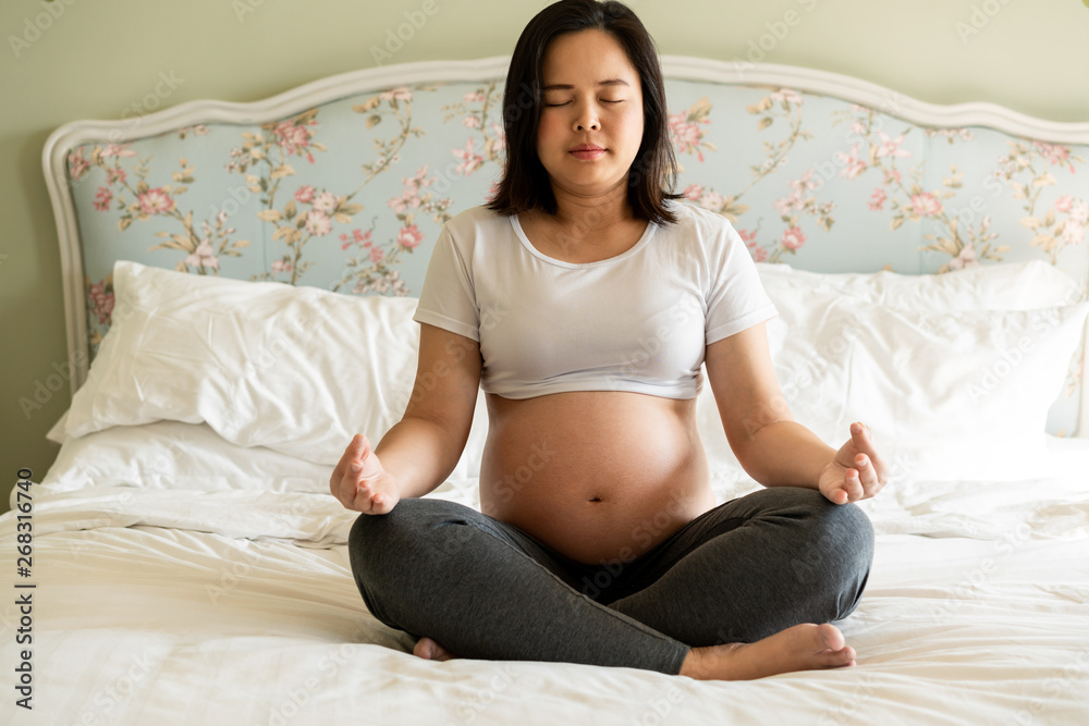 Pregnant woman doing yoga exercise on bed in bedroom at home while taking care of her child. The hap