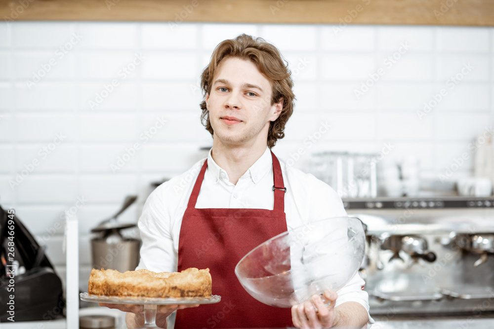 Portrait of a handsome young salesman in red apron standing with fresh pie at the modern pastry shop