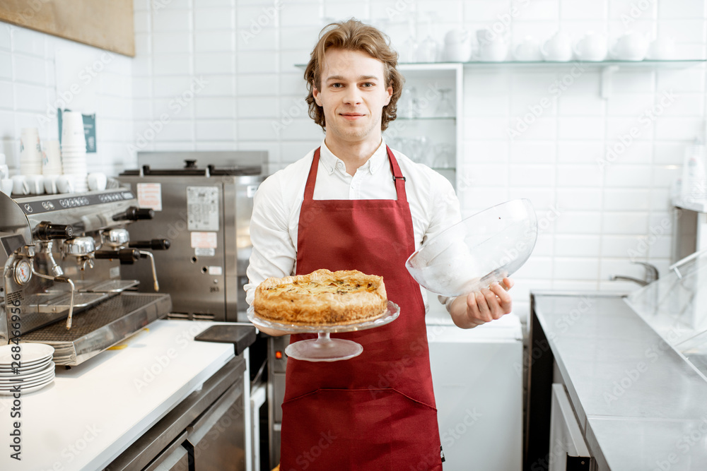 Portrait of a handsome young salesman in red apron standing with fresh pie at the modern pastry shop