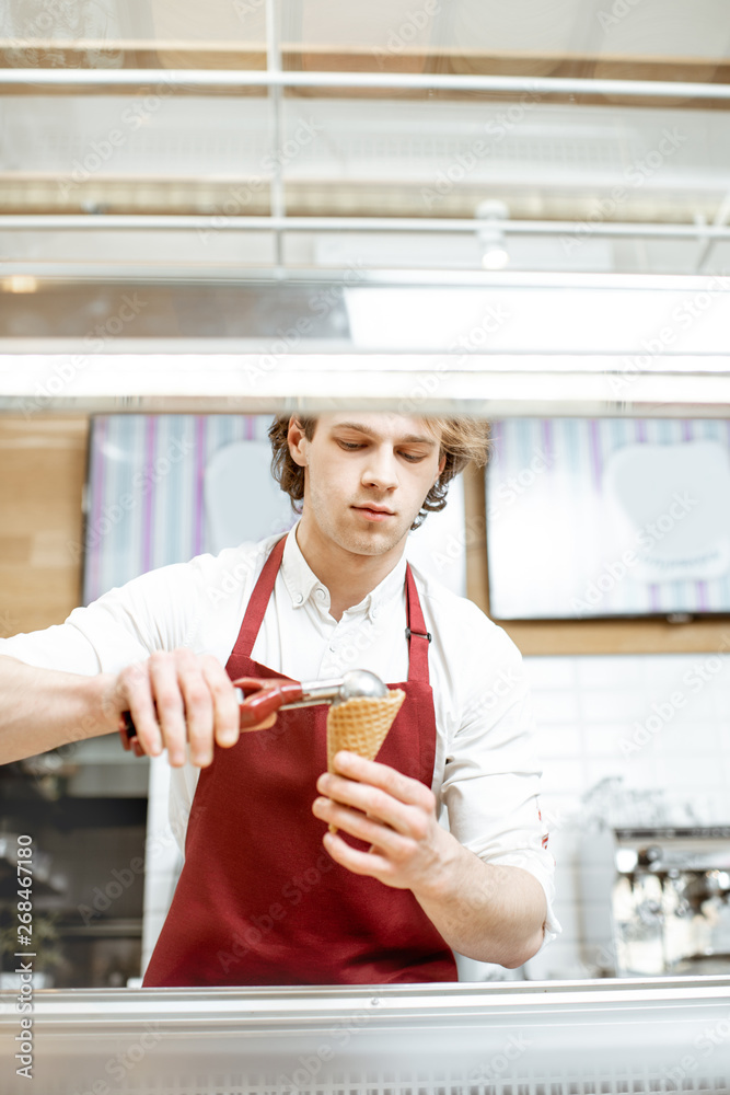 Young salesman putting ice cream ball into the waffle cone with professional tool at the pastry shop