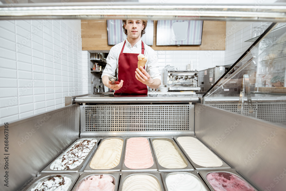 Salesman making ice cream in the waffle cone while standing near the ice cream trays at the pastry s