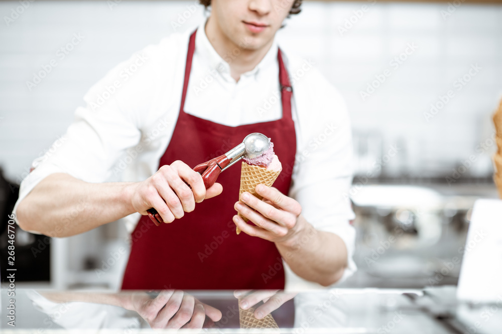 Handsome salesman in red apron making ice cream in waffle cone for selling in the modern pastry shop