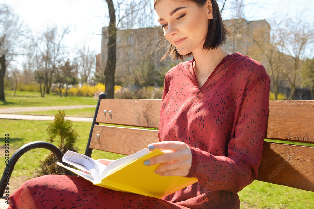 Beautiful young woman reading book on bench in park