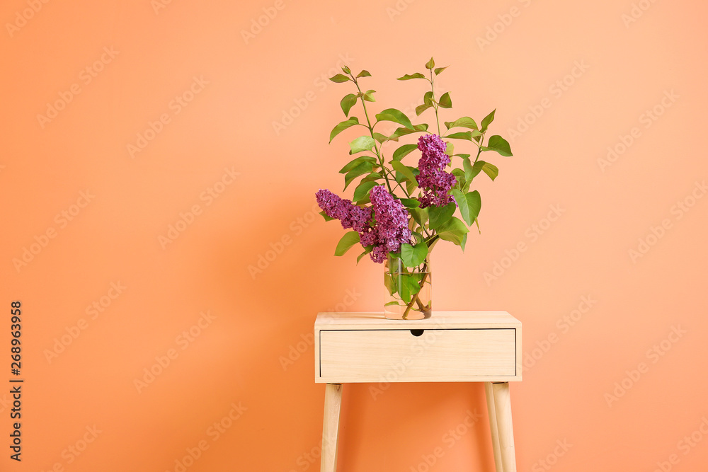 Beautiful lilac flowers in vase on table against color background