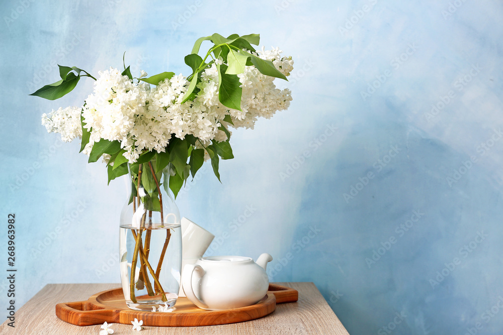 Beautiful lilac flowers in vase with teapot on table against color background