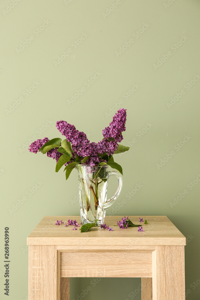 Beautiful lilac flowers in jug on table against color background