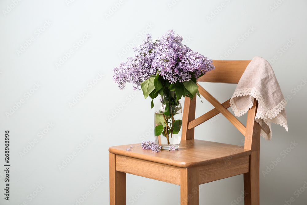 Beautiful lilac flowers in vase on chair against light background