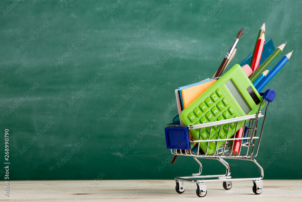 Education concept - school supplies in a shopping cart on the desk in the auditorium, blackboard bac