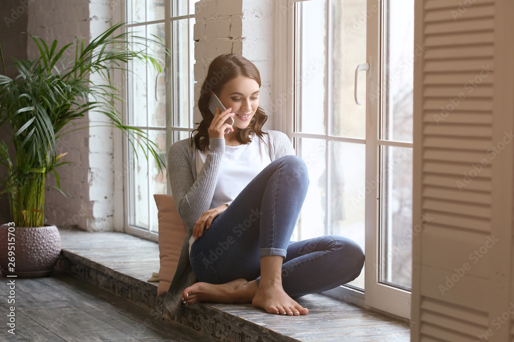 Beautiful young woman talking by mobile phone near window at home