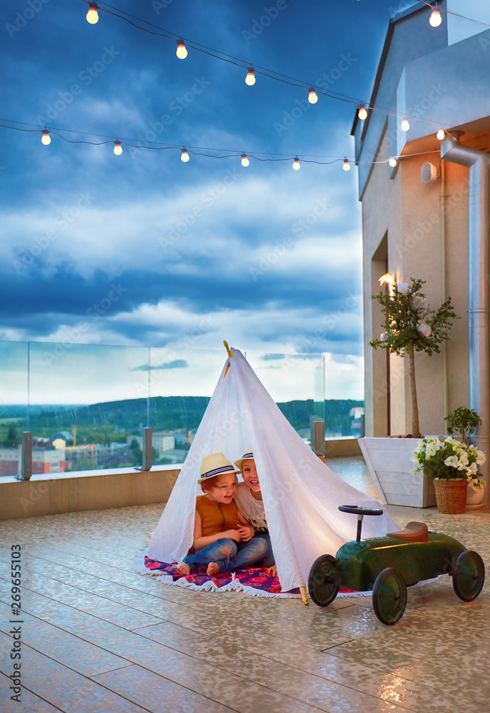 happy kids are hiding in handmade wigwam from a sudden summer rain at the patio, playing together