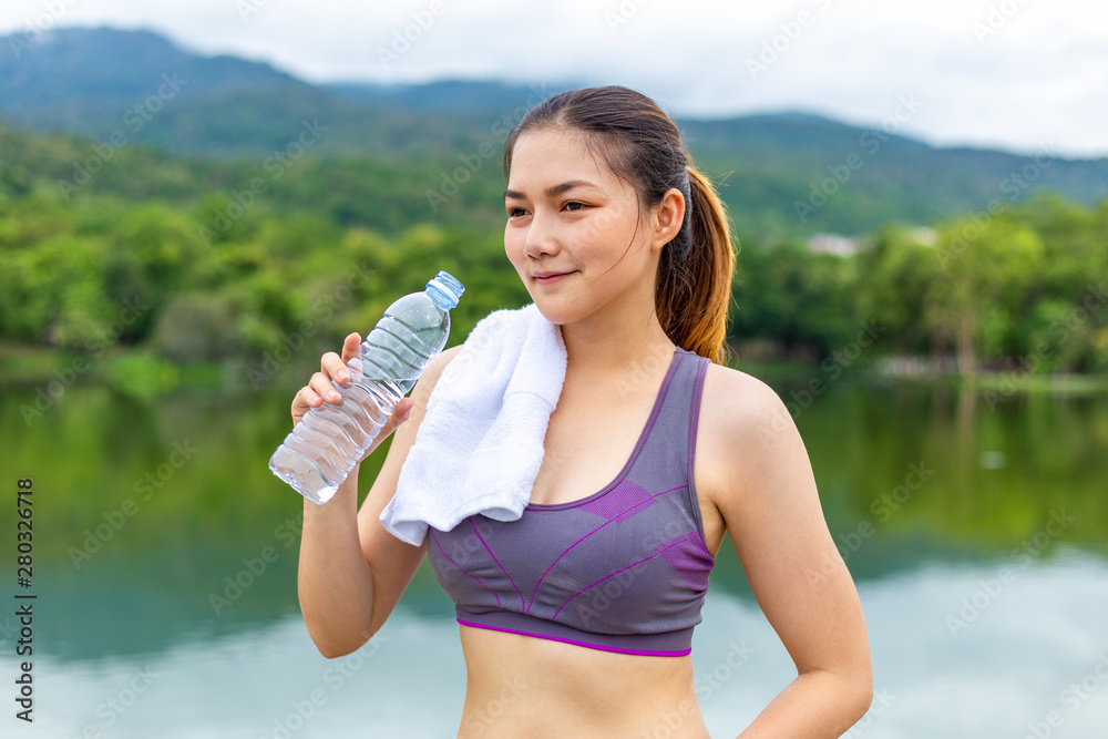 Beautiful Asian athlete woman drinking water during her break from morning  exercise at a lake park 