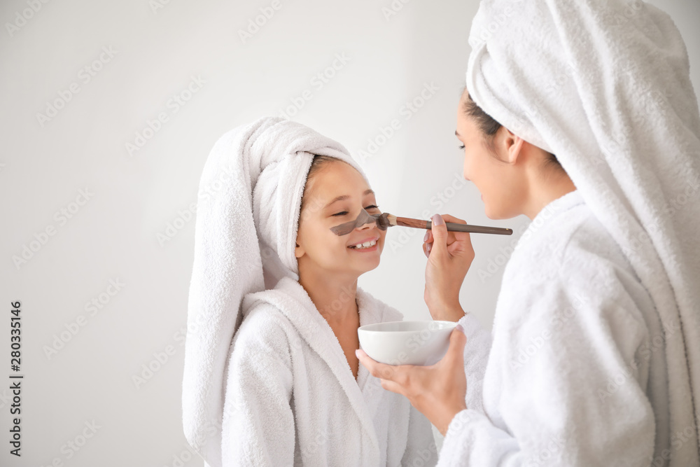 Mother applying mask onto face of her little daughter against light background