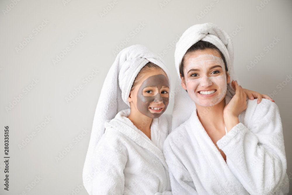 Mother and her little daughter in bathrobes and with facial masks against light background