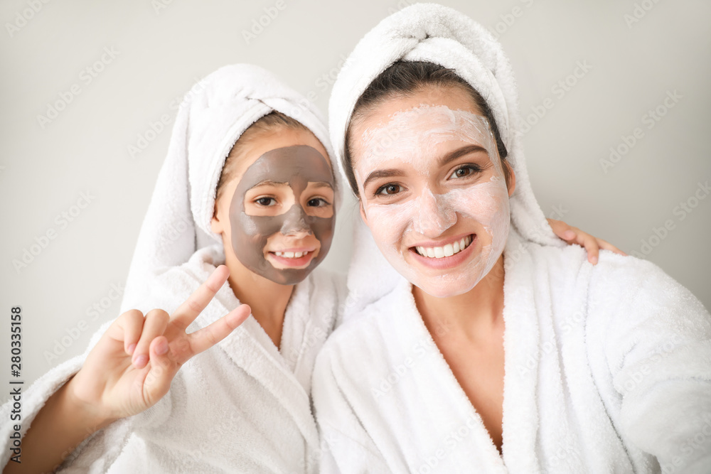 Mother and her little daughter in bathrobes and with facial masks against light background