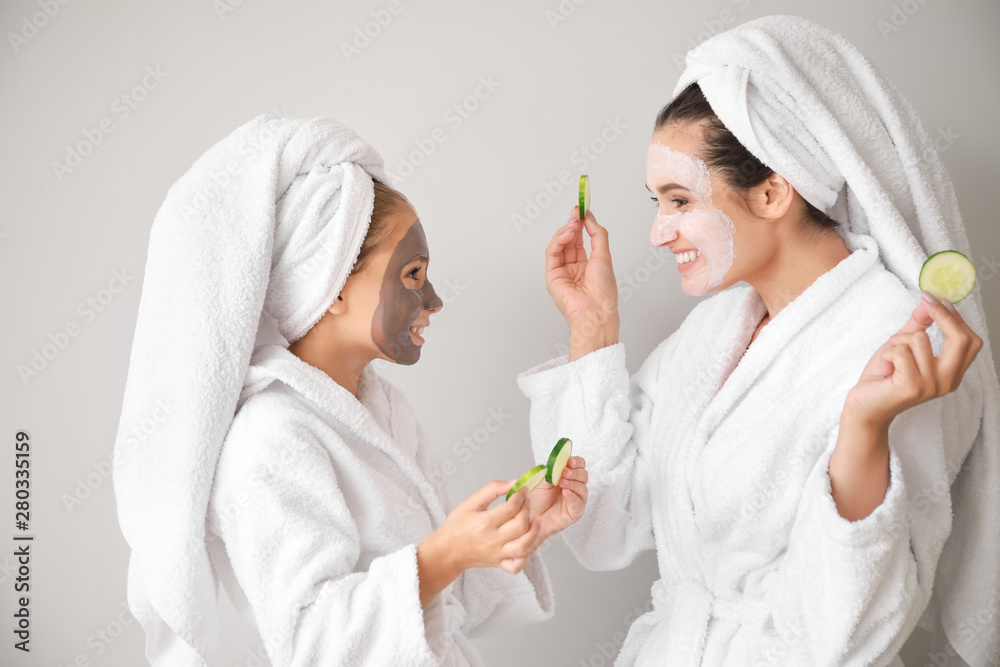 Mother and her little daughter with facial masks and cucumber slices on light background