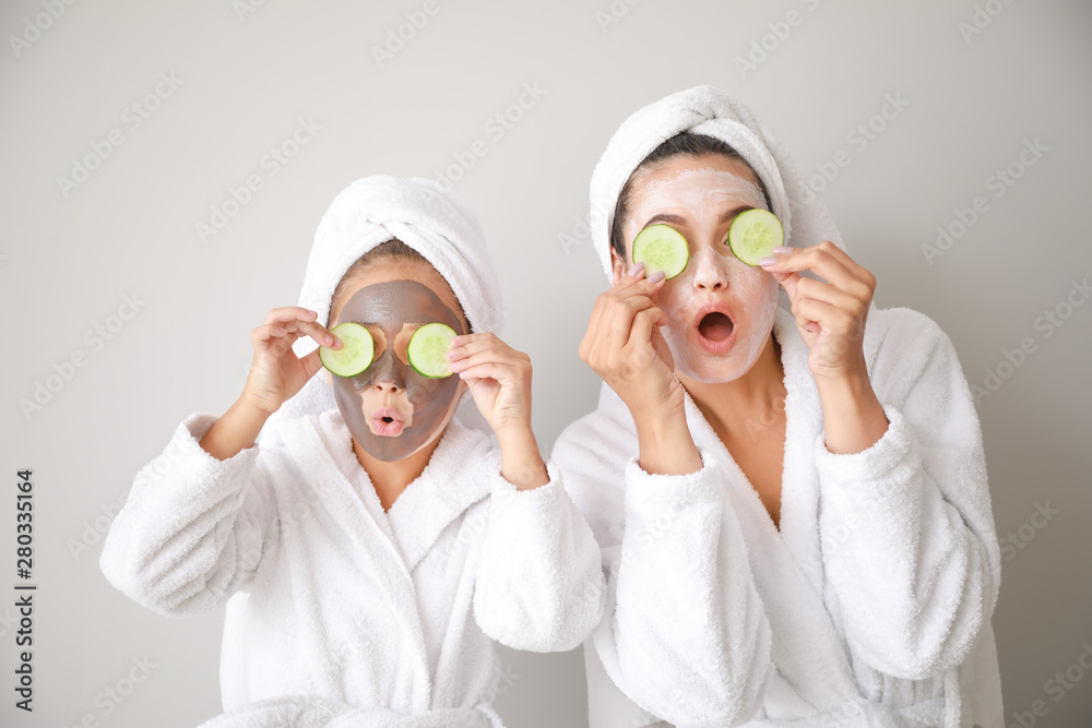 Funny mother and her little daughter with facial masks and cucumber slices on light background