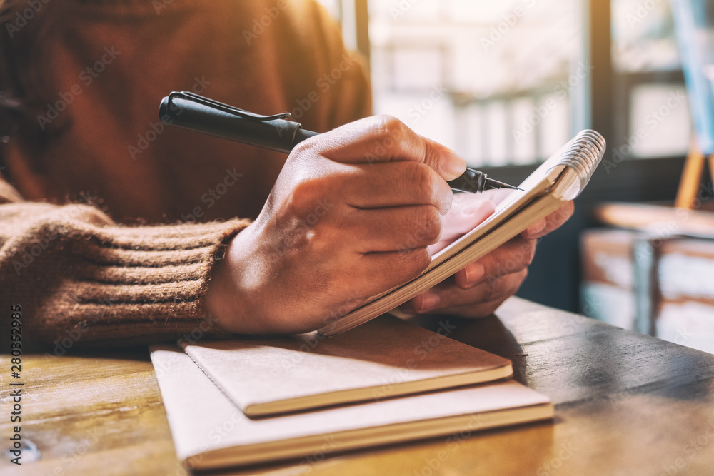 Closeup image of a woman holding and writing on blank notebook with fountain pen on wooden table