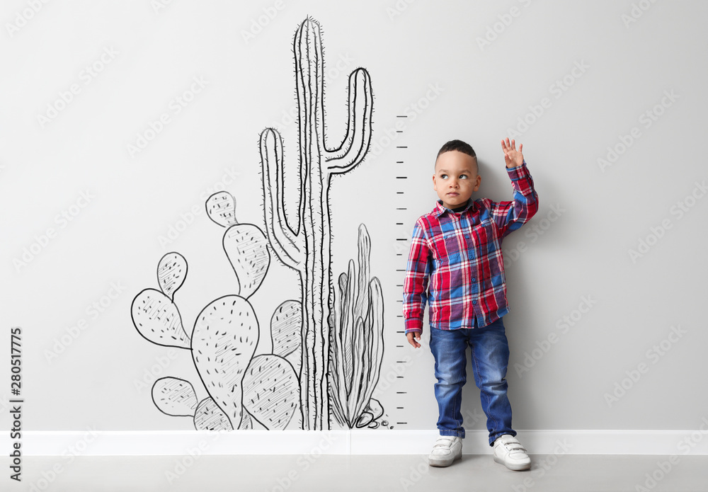 Cute little boy measuring height near light wall with drawn cacti