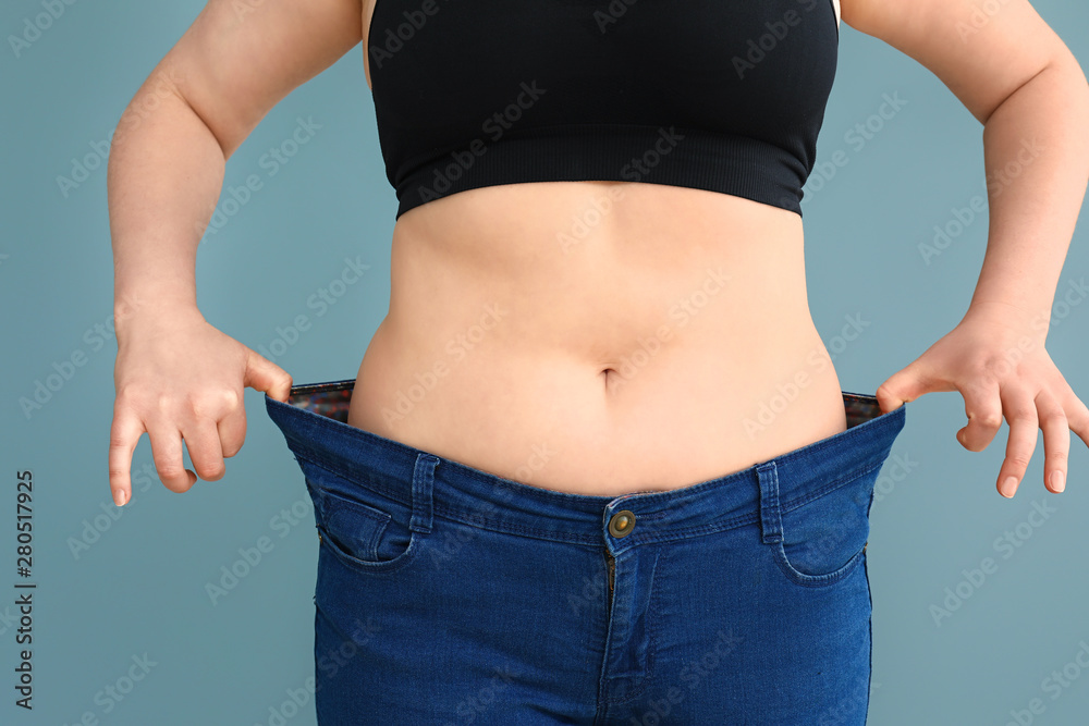 Overweight woman in loose jeans on color background. Weight loss concept