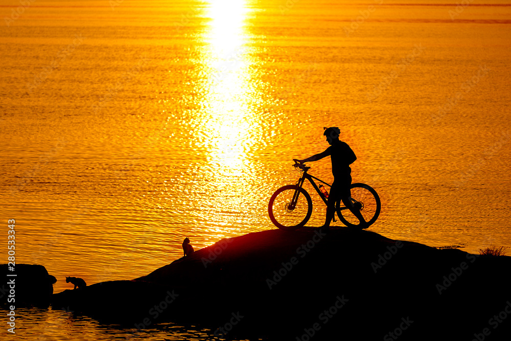 Silhouette of sportsman riding a bicycle on the beach. Colorful sunset cloudy sky in background.