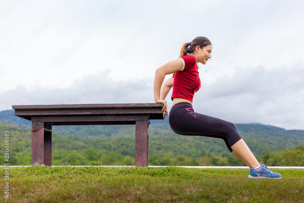 Beautiful young Asian woman doing tricep dips exercise outdoor at a park, cloudy sky and mountains b