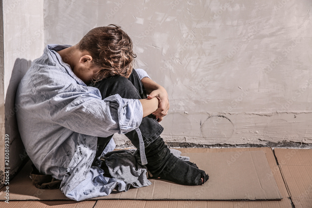 Homeless little boy sitting on floor near wall