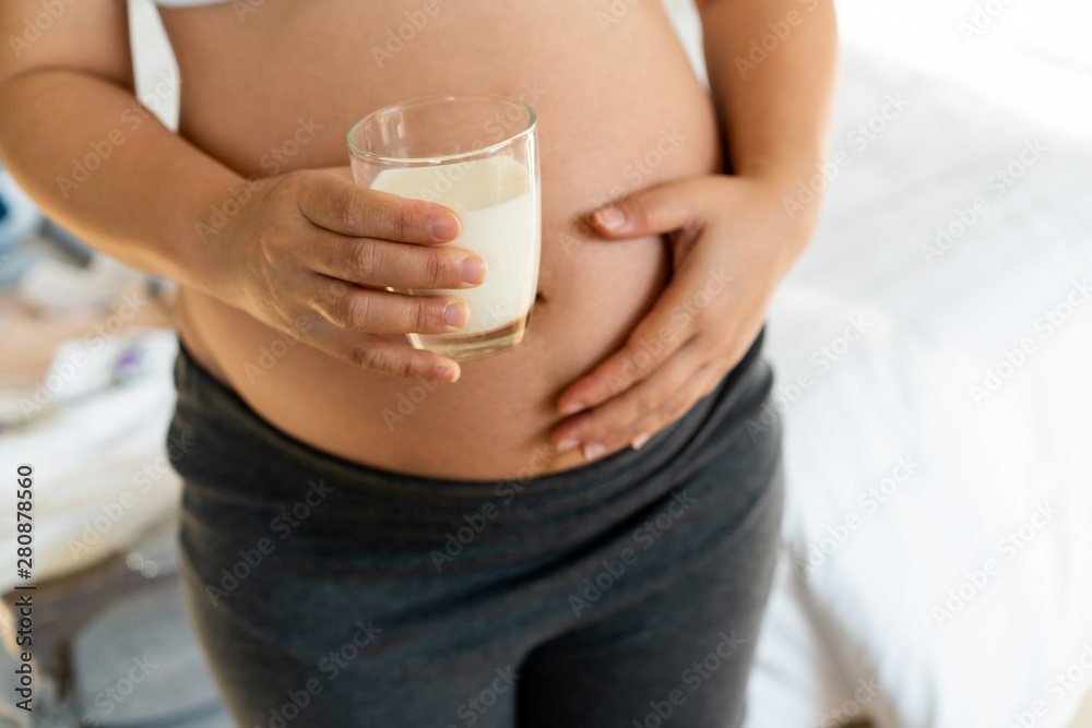 Happy pregnant woman drinks milk in glass at home while taking care of her child. The young expectin