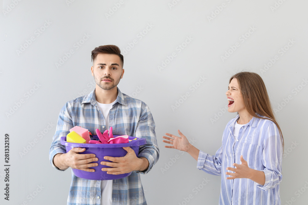 Angry wife scolding her sad husband with cleaning supplies on grey background