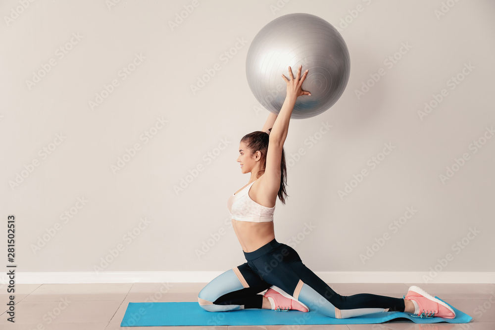 Young sporty woman with fitball doing exercises near light wall