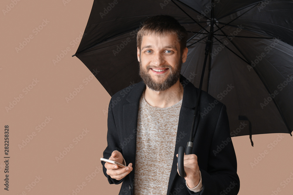 Handsome man with umbrella and mobile phone on color background