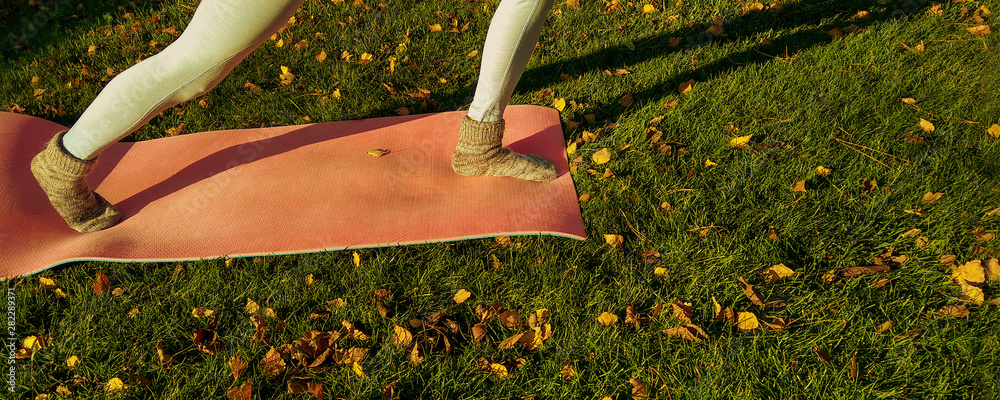 Yoga girl in autumn park with yoga mat on green carpet with fallen yellow leaves. Funny autumn legs 