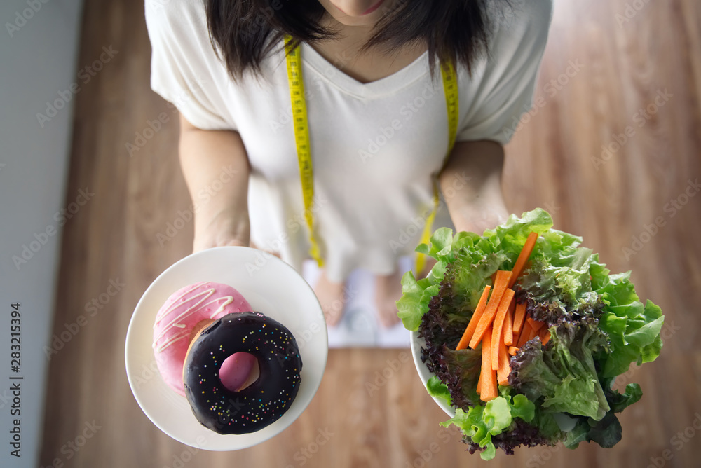 年轻的亚洲女孩拿着蔬菜沙拉和甜甜圈站在体重秤上的俯视图