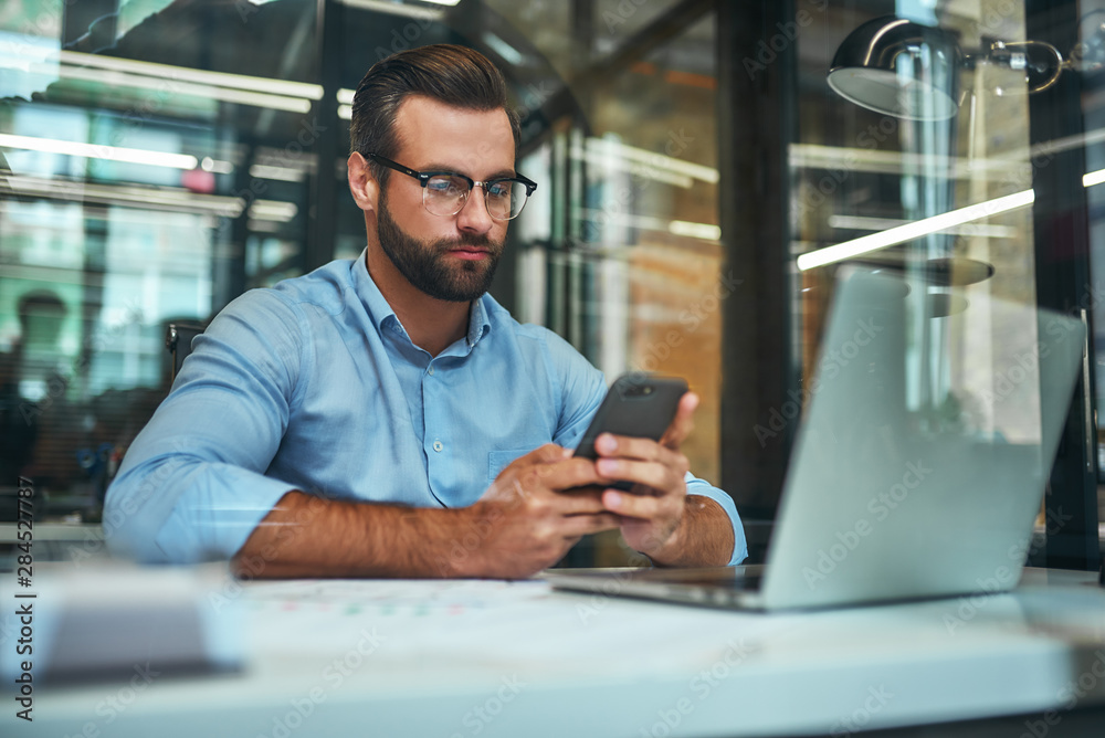 Important call. Portrait of young focused businessman in eyeglasses and formal wear looking at his s