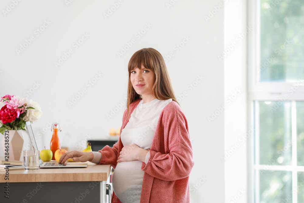 Beautiful pregnant woman with laptop in kitchen