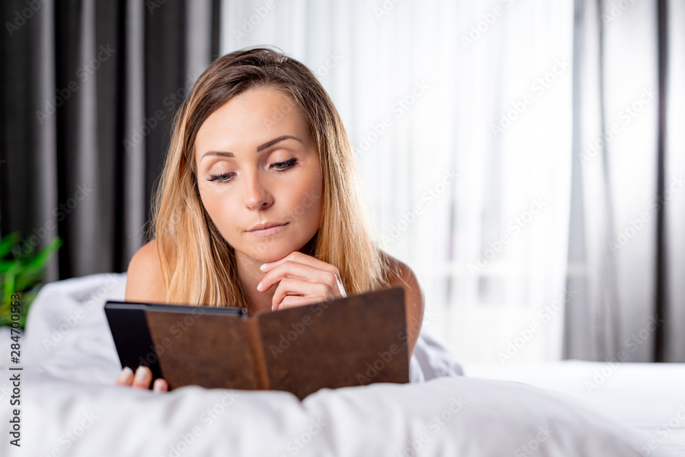 Relaxed pretty woman reading book using ebook reader or tablet lying on bed