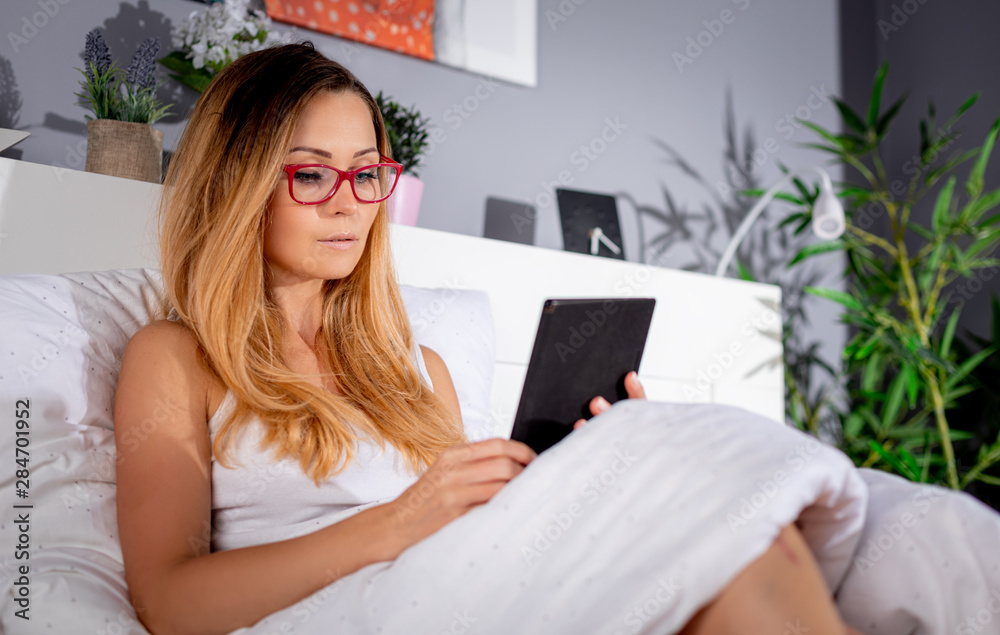 Beautiful young woman in glasses reading book using ebook reader or tablet lying on bed