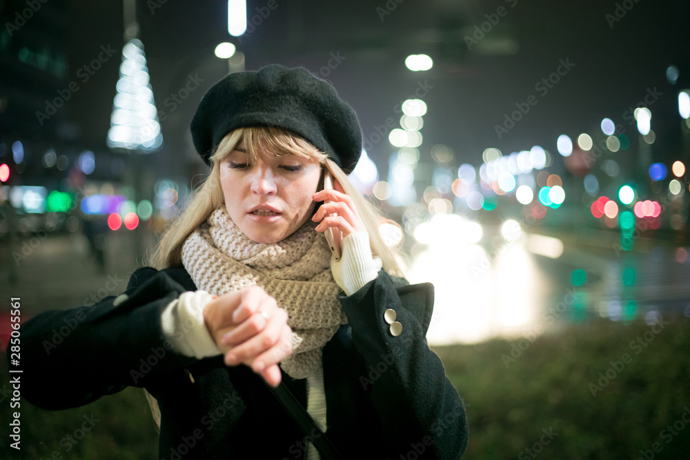 Pretty young stylish woman walking in the night city streets using modern smartpone, bokeh illuminat