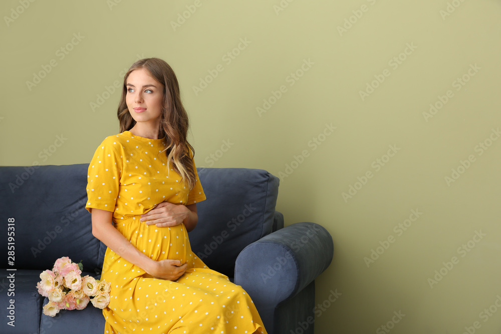 Beautiful pregnant woman with bouquet of flowers sitting on sofa against color background