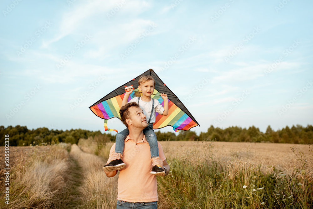 一位幸福的父亲和年幼的儿子的画像，肩上放着五颜六色的空中风筝，在蓝天上