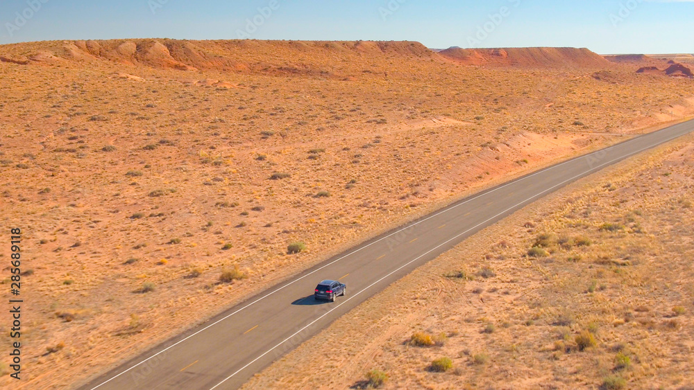 AERIAL: Black SUV jeep driving along the straight empty road through dry desert