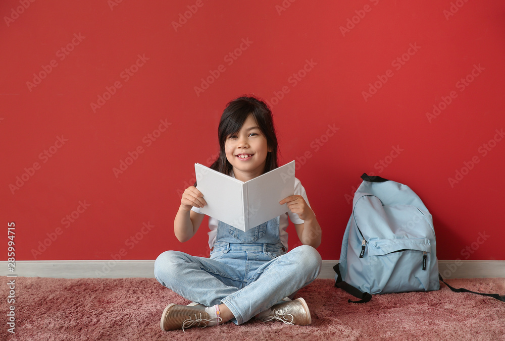 Portrait of little schoolgirl reading book near color wall