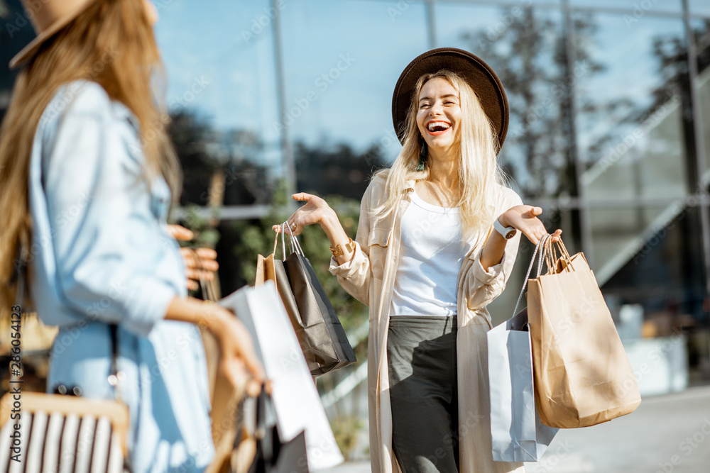 Two happy girlfriends walking with shopping bags in front of the shopping mall, feeling satisfied wi