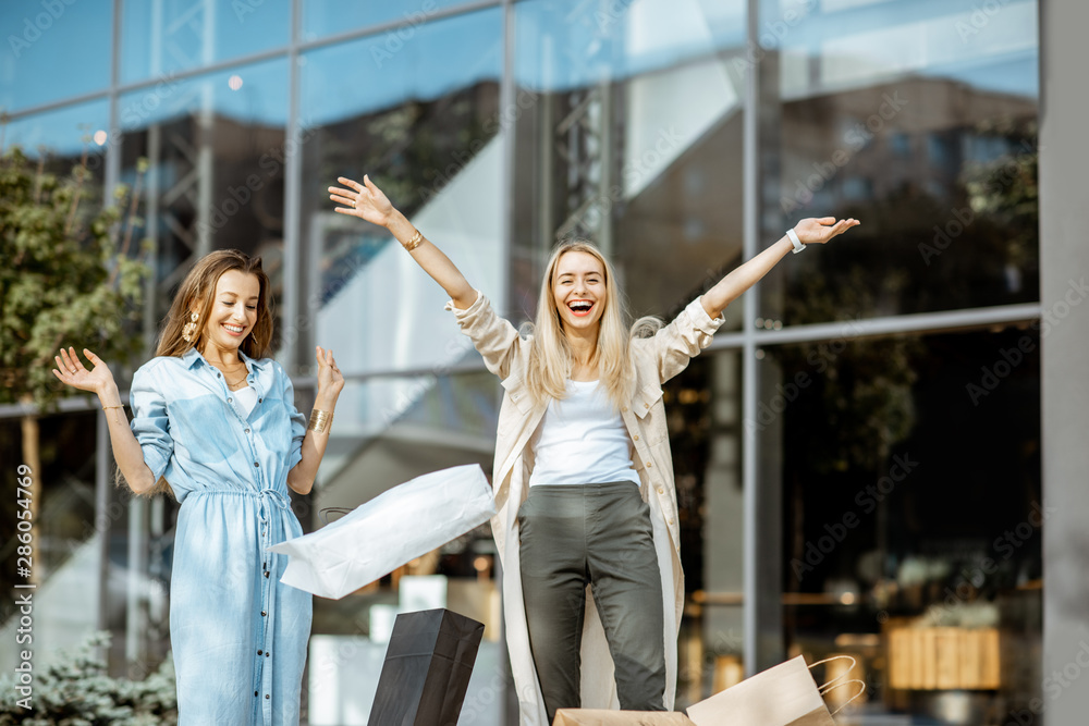 Two happy girlfriends throwing up shopping bags in front of the shopping mall outdoors, feeling exci