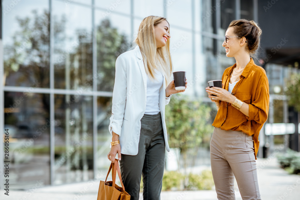 Two young businesswomen talking near the office building, having a small talk during the coffee brea