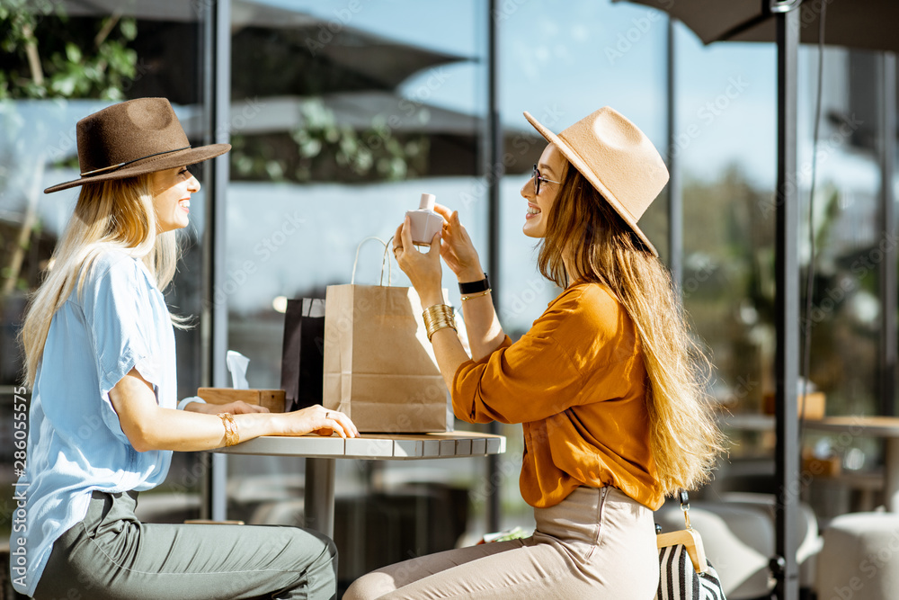 Two stylish girlfriends sitting at the modern cafe terrace outdoors, looking on a new purchases afte