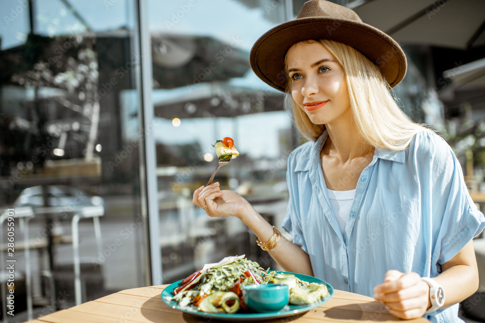 Stylish young woman eating healthy salad on a restaurant terrace, feeling happy on a summer day