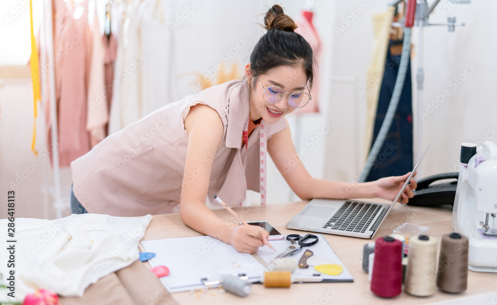 Beautiful asian woman fashion designer tailor fabric working and sewing her sketch clothes with lapt