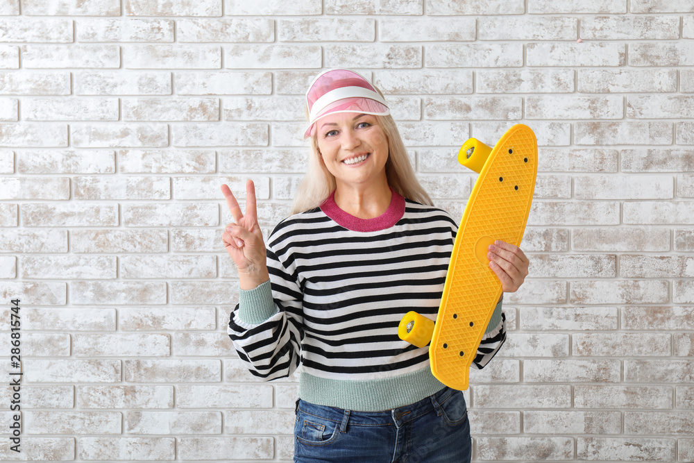 Stylish mature woman with skateboard showing victory gesture on brick background